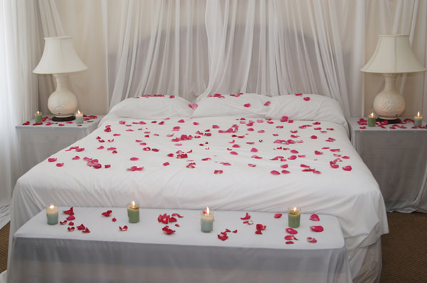 اتاق عروسی که روی روتختی سفید آن گلبرگ های صورتی رنگ پخش شده است و روی میزهای کنار تختی و صندلی پایین تخت آن شمع های روشن قرار دارد